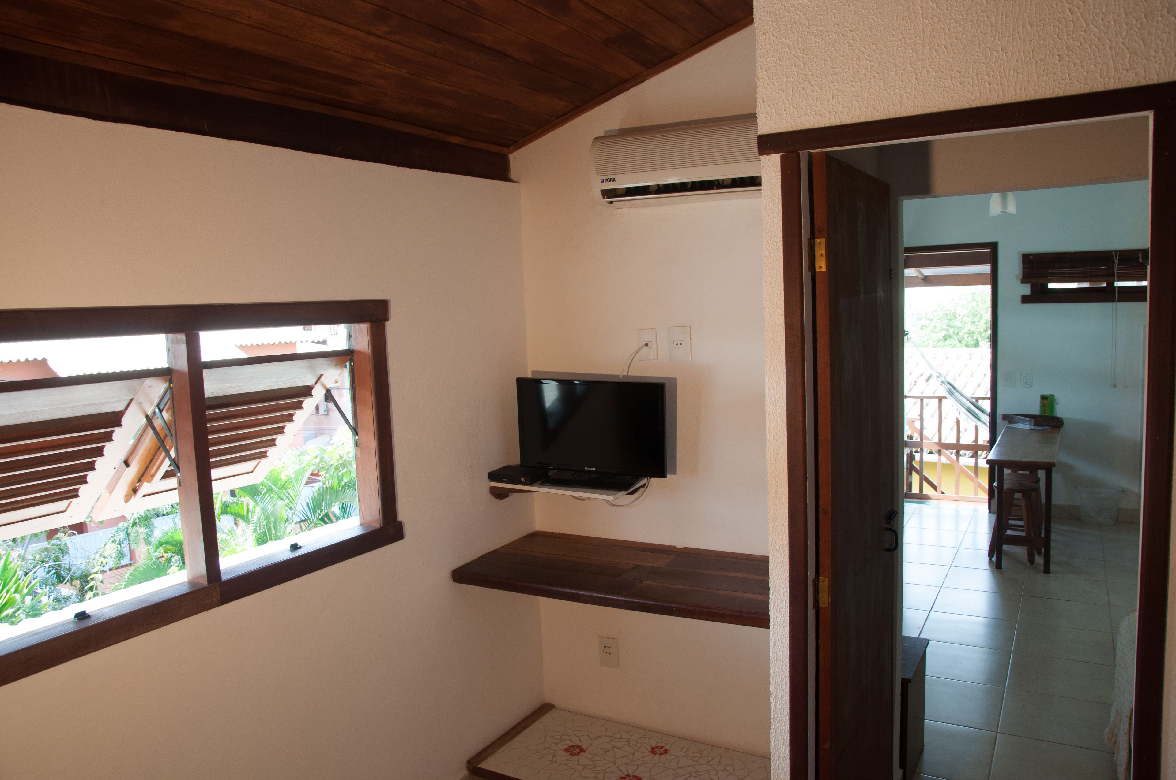 Ein Zimmer mit an der Wand befestigtem Fernseher, einem Fenster mit Blick auf Grünflächen und einer Holzbalkendecke.