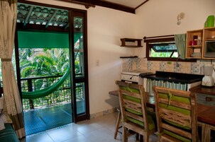 Una cocina con vistas a exuberante follaje a través de una puerta de vidrio, una zona de comedor con sillas de madera y un pequeño estante con utensilios de cocina.