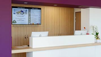 A hotel reception desk with a digital display board above it, a wooden cabinet to the left, and a potted orchid on the right.
