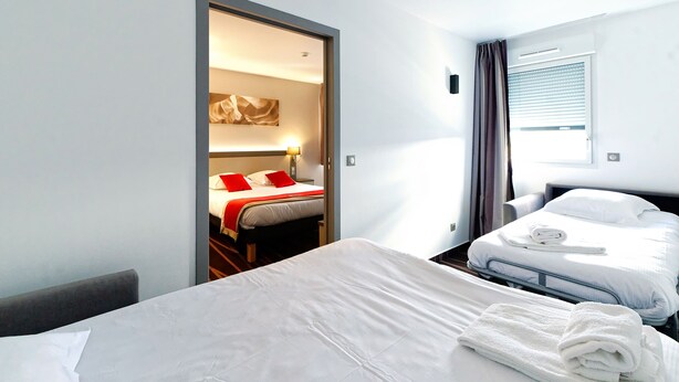 A hotel room with two beds, one with red pillows, a large window with blinds, and a door leading to another room. - Hôtel Akena Besançon (Chemaudin)