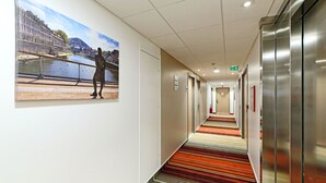 A hotel corridor with a colorful striped carpet, a painting of a person by a river, and a modern elevator. - Hôtel Akena Besançon (Chemaudin)