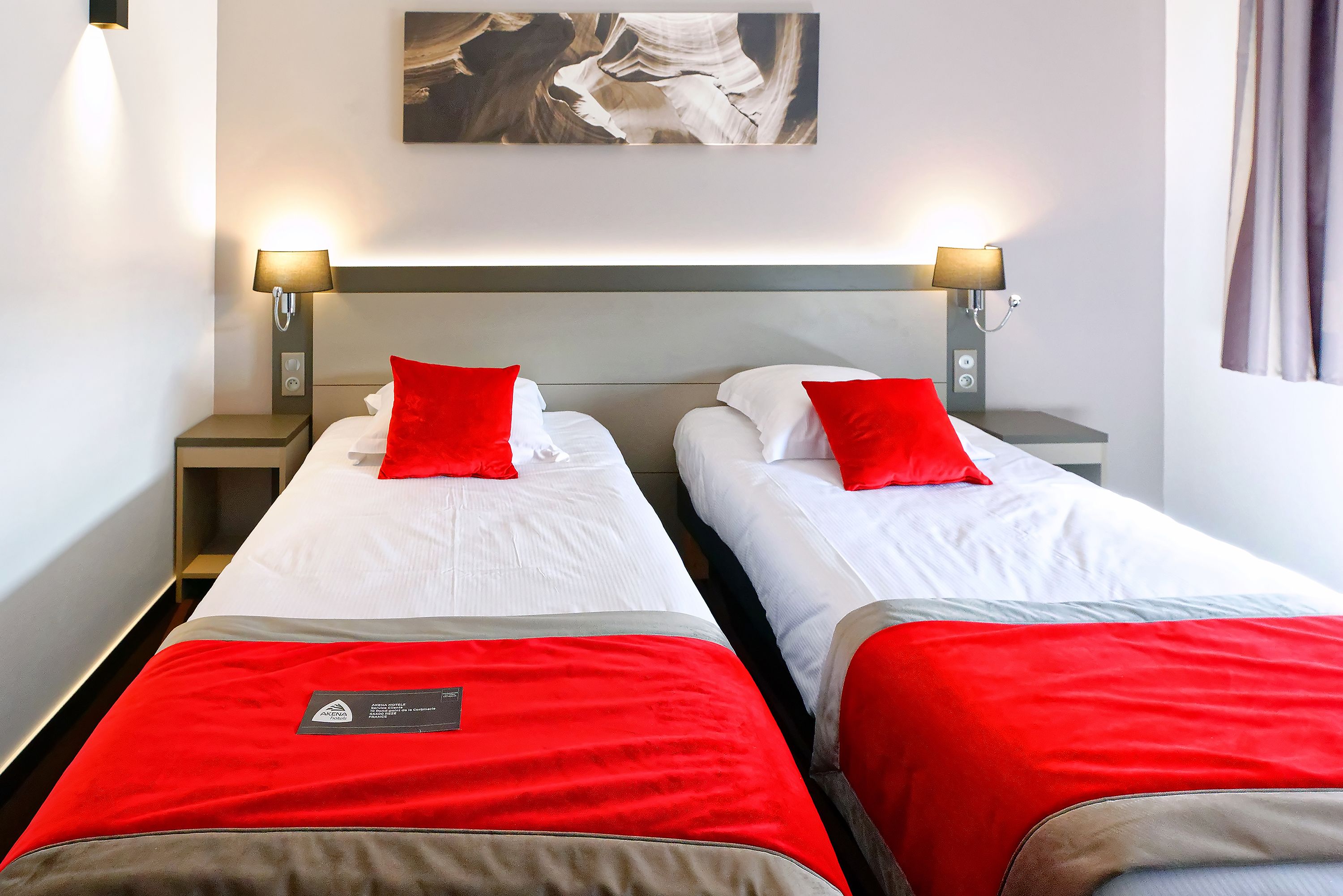 Une chambre d’hôtel avec deux lits, chacun agrémenté d’un oreiller rouge et d’une couvre-lit blanche. De chaque côté des lits, on trouve des lampes fixées au mur, et un tableau est accroché au-dessus.