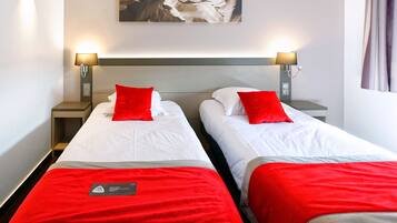 Une chambre d’hôtel avec deux lits, chacun agrémenté d’un oreiller rouge et d’une couvre-lit blanche. De chaque côté des lits, on trouve des lampes fixées au mur, et un tableau est accroché au-dessus.