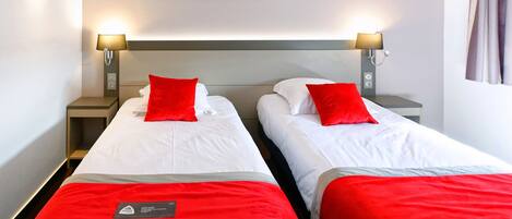 A hotel room with two beds, each with a red pillow and a white bedspread. There are wall-mounted lamps on either side of the beds, and a painting above them.