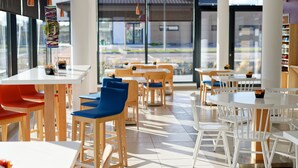A modern restaurant interior with white and wooden tables and chairs, large windows, and pendant lighting. - Hôtel Akena Besançon (Chemaudin)