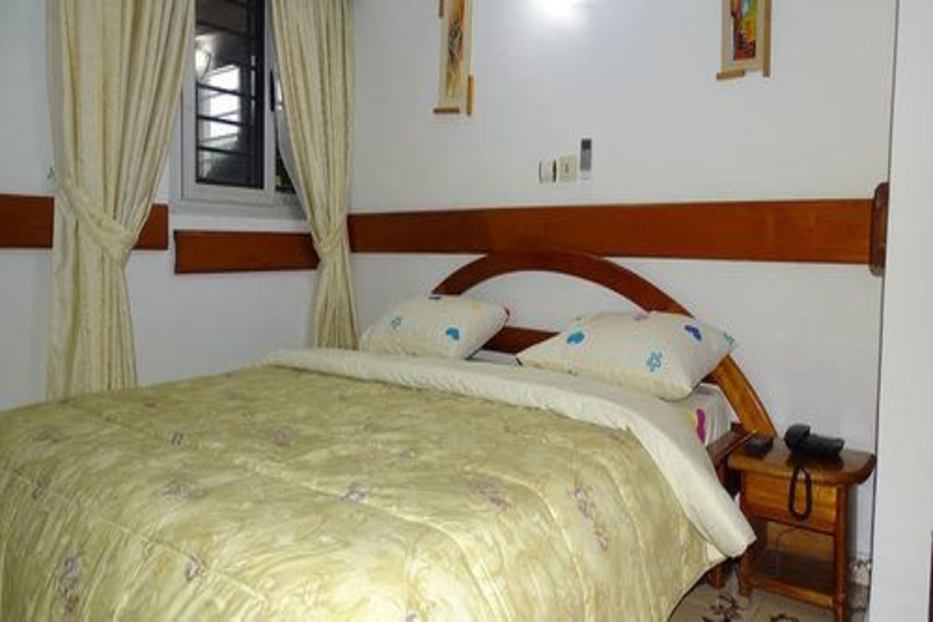 Un dormitorio con cama, cortinas, cabecero de madera, mesita de noche con lámpara y teléfono fijo en la pared.