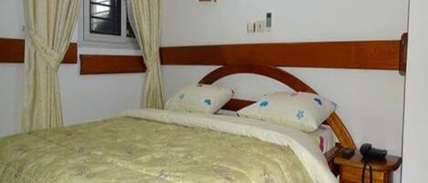Un dormitorio con cama, cortinas, cabecero de madera, mesita de noche con lámpara y teléfono fijo en la pared.