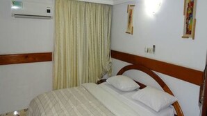 A hotel room with a wooden headboard, a bed with white linens, a window with curtains, and an air conditioning unit on the wall. - Residence Hotel Badjo (Abidjan)