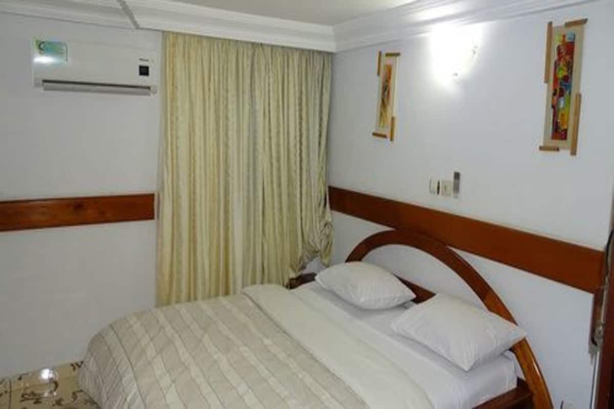 A hotel room with a wooden headboard, a bed with white linens, a window with curtains, and an air conditioning unit on the wall.