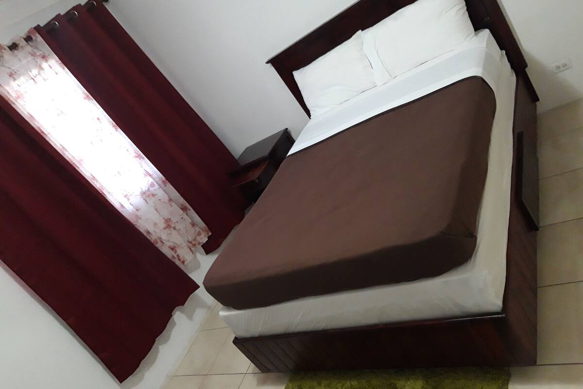 A bed with a brown mattress and white pillows, a wooden headboard, and a green rug on a tiled floor.