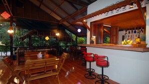 Um restaurante com mesas e cadeiras de madeira, um espaço de bar com banquetas e vista para o exterior com vegetação.