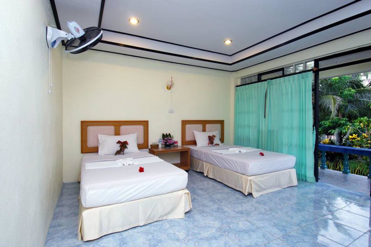 A hotel room with two beds, a ceiling fan, a small table with a vase, and a view of greenery through a window with a curtain.