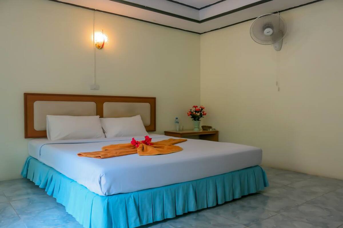 A neatly made bed with a blue skirt, a wooden headboard, and a wall-mounted fan.
