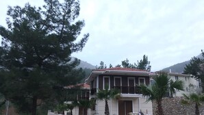 A two-story house with a stone wall and a red-tiled roof, surrounded by trees and a mountainous backdrop. - Likya Lodge Adrasan (Kumluca)