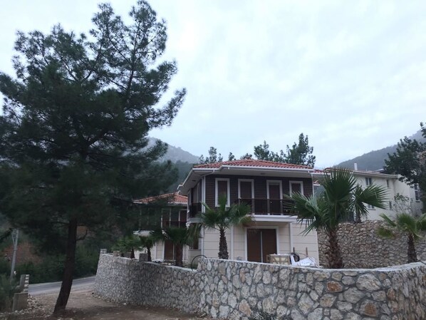 Uma casa de dois andares com muro de pedra e telhado de telhas vermelhas, cercada por árvores e com montanhas ao fundo.