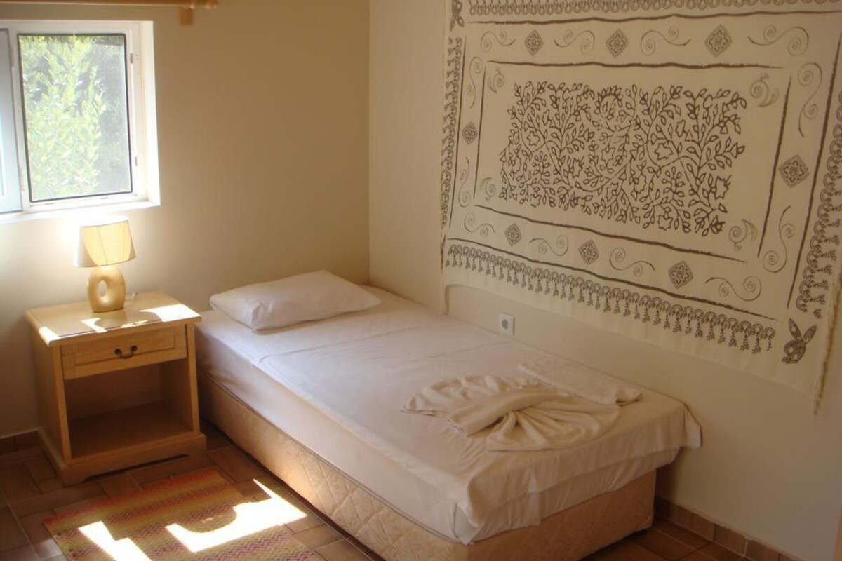 Uma cama de solteiro com colcha branca e toalha, uma mesa de cabeceira de madeira com abajur, e uma parede com padrão decorativo.