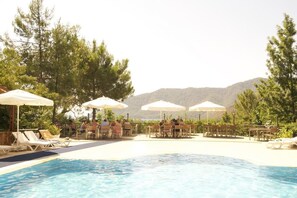 Área de piscina com espreguiçadeiras e guarda-sóis, cercada por árvores e vista para uma montanha.