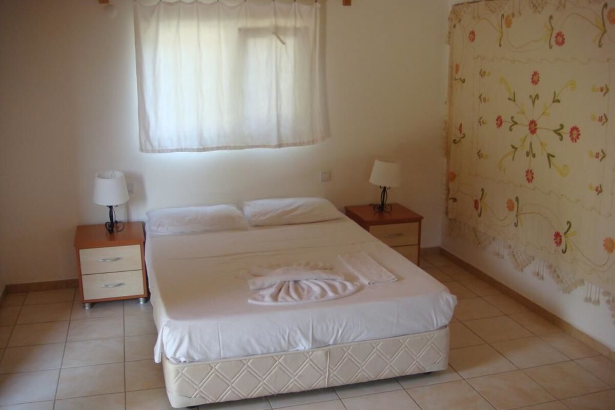 Quarto com uma cama grande, duas mesas de cabeceira com luminárias, uma janela com cortina translúcida e um papel de parede com padrão floral.