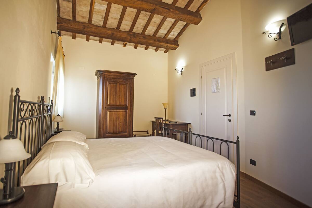 A bedroom with a wooden ceiling, a bed with white linens, a nightstand with a lamp, a desk with a chair, and a wardrobe.