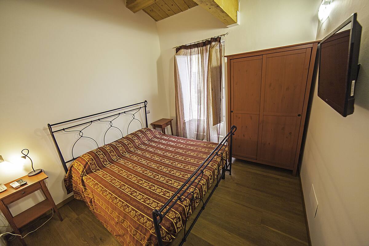 A bedroom with a bed, a nightstand, a wooden wardrobe, and a wall-mounted TV.