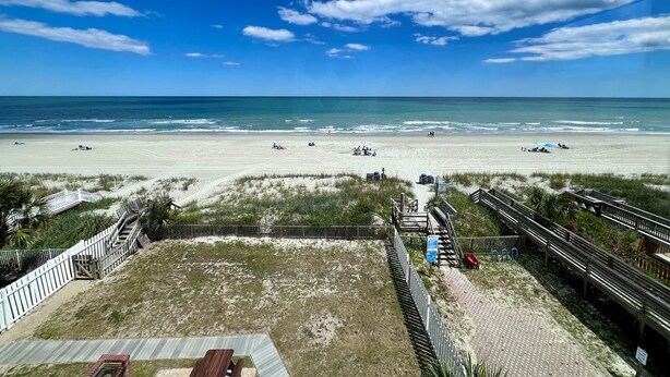 A beach with a wooden boardwalk leading to a grassy area with a bench.