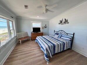 A bedroom with a bed, a television, a dresser, a bench, and a window overlooking the beach.