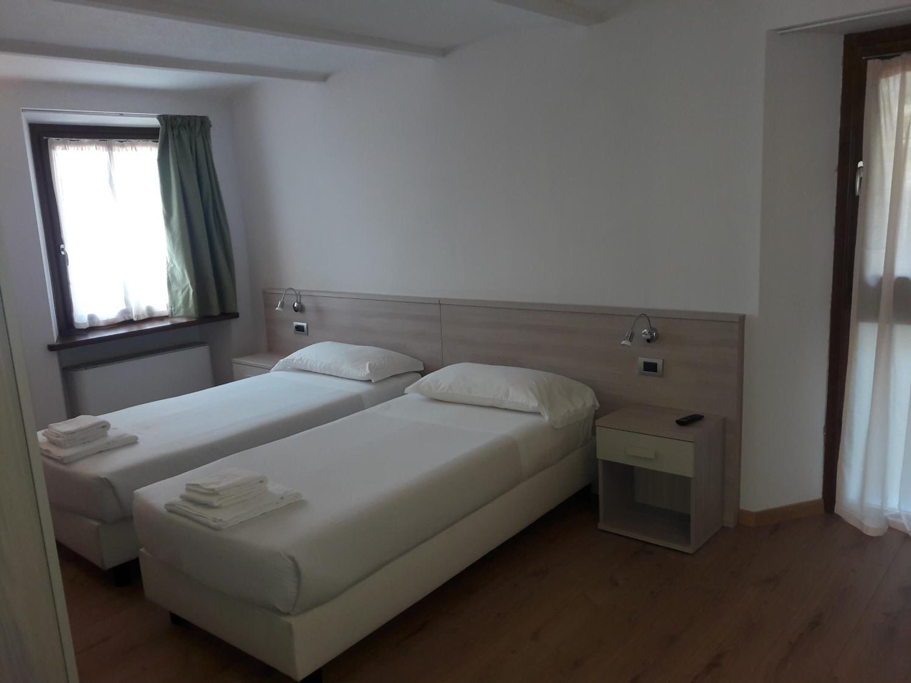 A hotel room with two single beds, a bedside table with a lamp, a window with curtains, and a door.