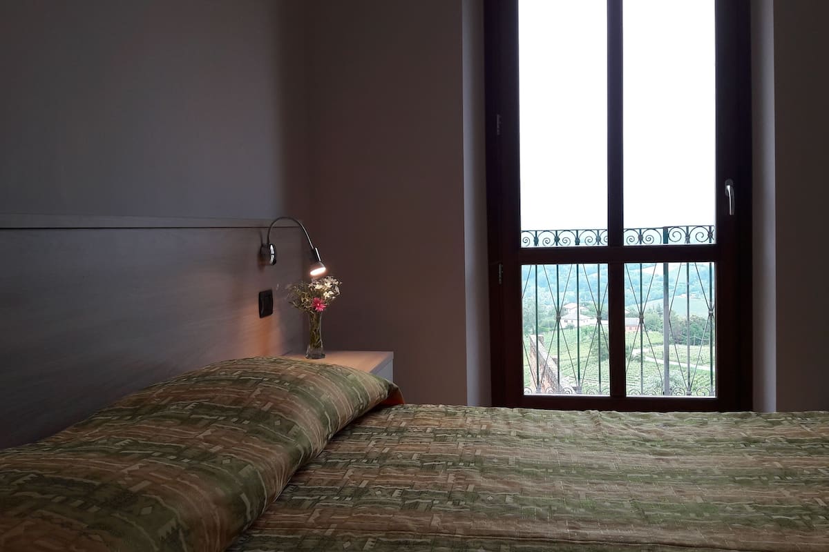 Ein Schlafzimmer mit einem Bett, einem Nachttisch, einer Lampe und einem Fenster mit Blick auf Grünflächen.