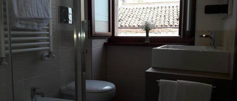 A bathroom with a bidet, toilet, and sink. There is a window with a view of a building with a bell tower.