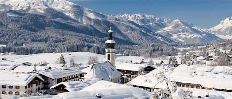 Ein schneebedecktes Dorf mit traditionellen Holzhäusern, einer markanten Kirche mit hohem Kirchturm und einem Berg im Hintergrund.