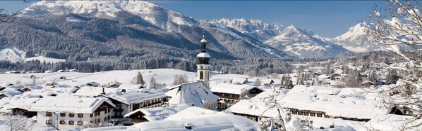 A snow-covered village with traditional wooden houses, a prominent church with a tall steeple, and a mountain backdrop.