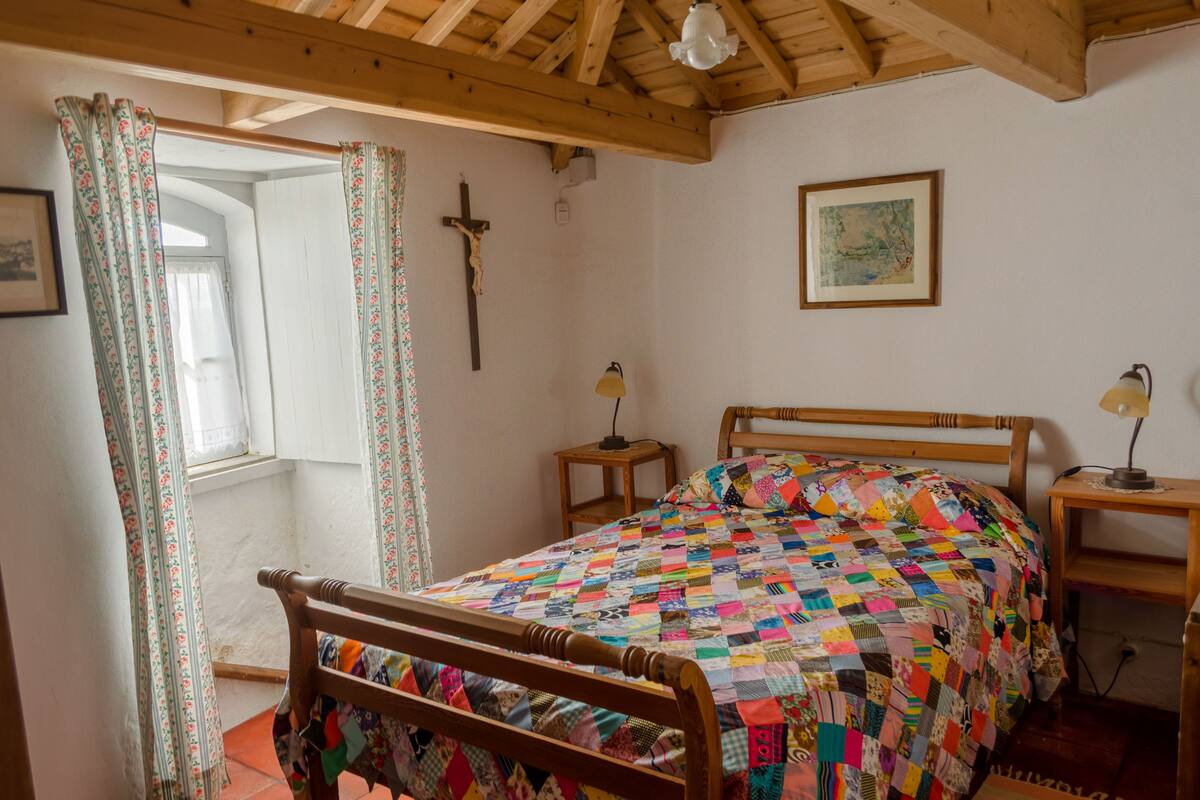 Une chambre à coucher avec un lit en bois, une croix accrochée au mur, une table de chevet avec une lampe et une fenêtre avec des rideaux.