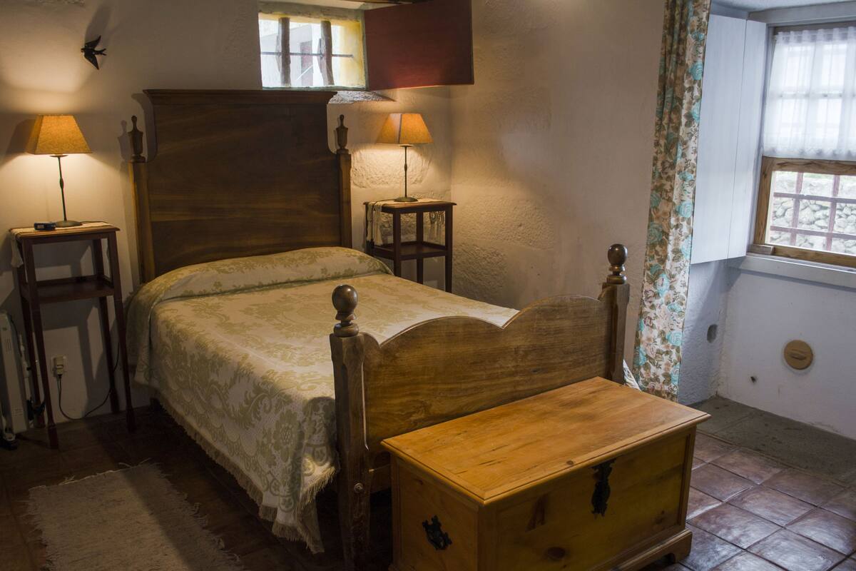 Un lit traditionnel en bois, doté d’une tête de lit, de deux tables de chevet avec des lampes, d’une commode en bois et d’une fenêtre avec des rideaux.