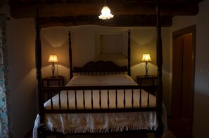 Un lit à baldaquin avec deux tables de chevet, chacune dotée d’une lampe, dans une pièce aux poutres apparentes et au décor simple et rustique.