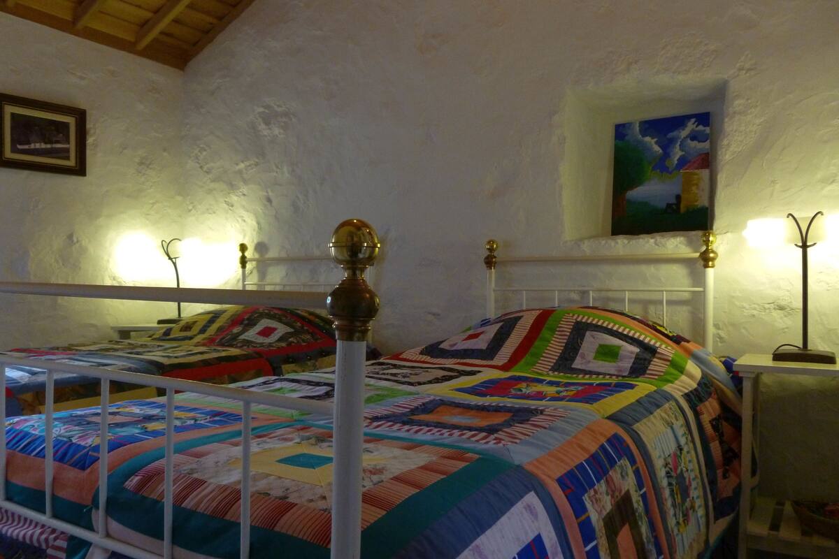 Une chambre à coucher avec deux lits, chacun doté d’un cadre blanc et d’une couverture colorée. On y trouve des tables de chevet avec des lampes, un tableau au mur et un plafond en bois.