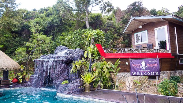 A pool with a waterfall, surrounded by lush greenery and a building with a 'Welcome' sign.