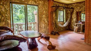 Quarto com parede de pedra, rede, mesa redonda e vista para o verde lá fora.