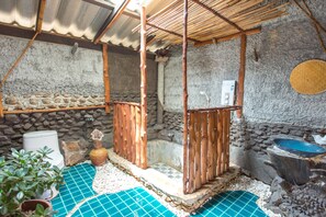 A rustic outdoor bathroom with a stone wall, a wooden shower enclosure, a blue tiled floor, and a stone basin.