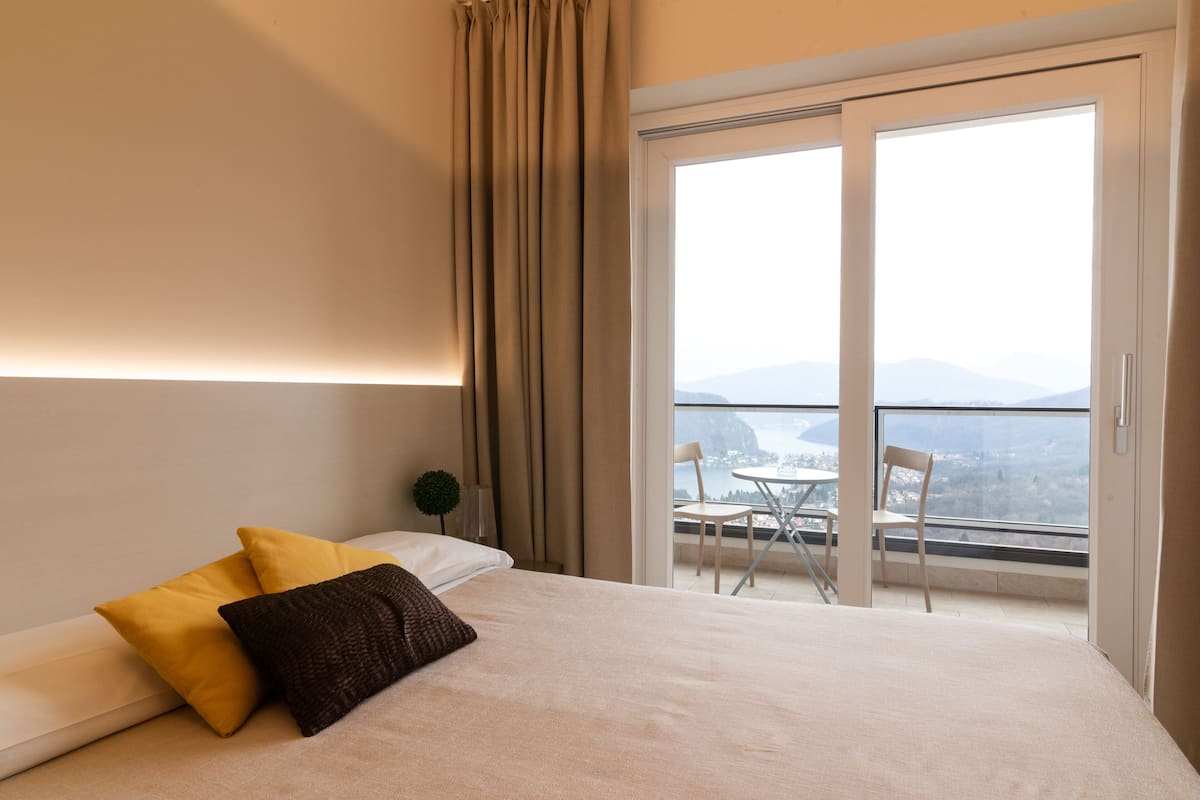 A hotel room with a bed, two pillows, a nightstand, a small plant, and a balcony with a table and chairs.