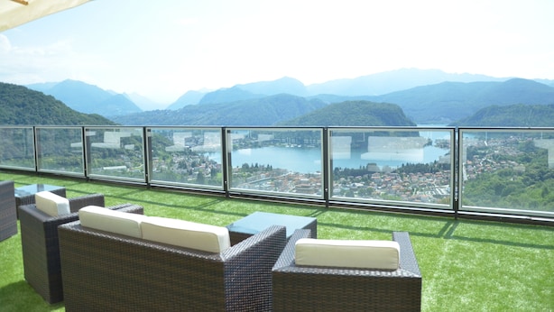 Terraza con mobiliario de mimbre y barandilla de vidrio con vistas a una ciudad y montañas.