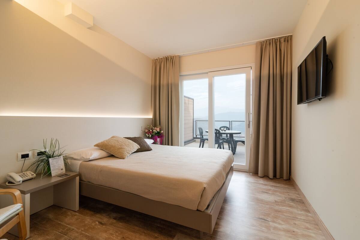 A hotel room with a bed, bedside table with a phone, a flat-screen TV, and a balcony with a table and chairs.