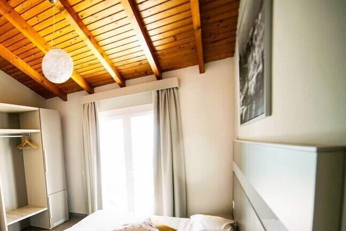 A bedroom with a wooden ceiling, a bed with white bedding, a wardrobe, and a window with curtains.