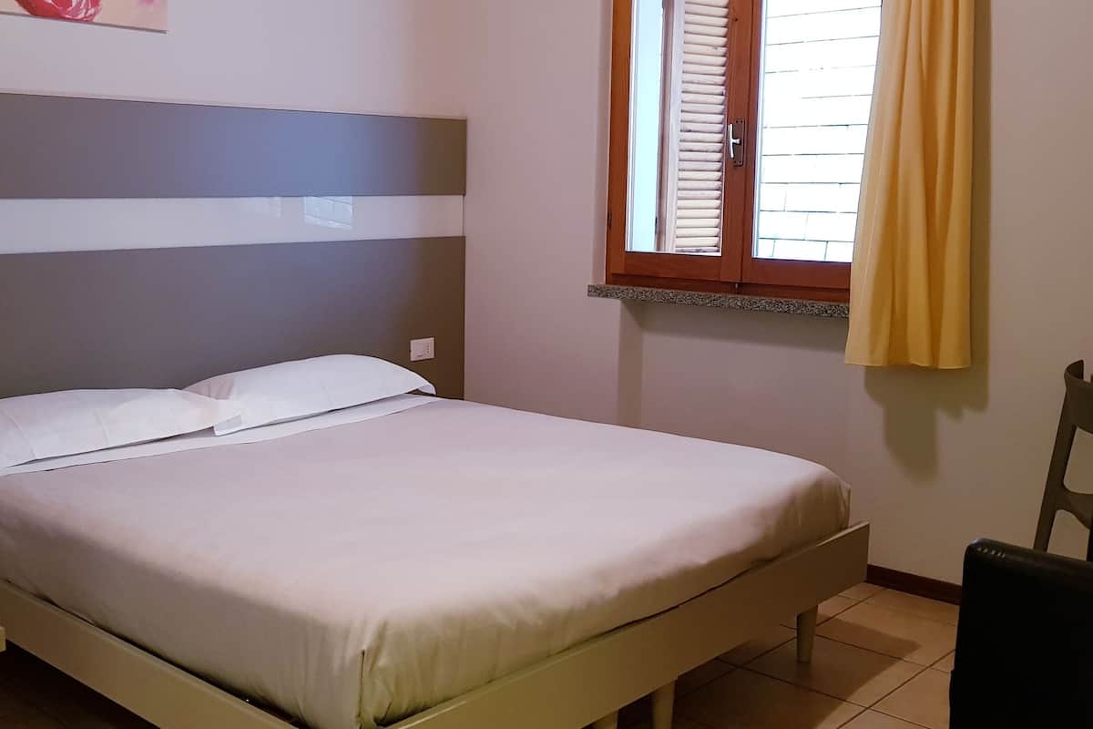 A single bed with a headboard, a bedside table, a chair, and a window with blinds.
