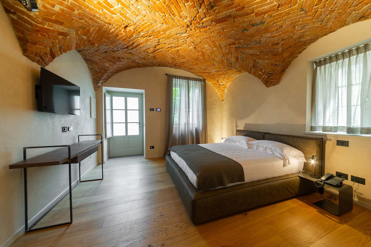 A bedroom with a brick ceiling, a bed with white bedding, a bedside table with a lamp, and a TV mounted on the wall.
