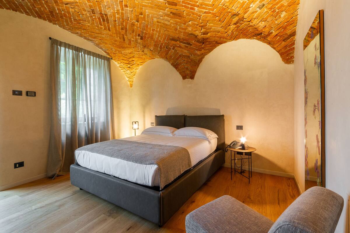A bedroom with a brick ceiling, a bed with a headboard, a bedside table with a lamp, and a window with curtains.