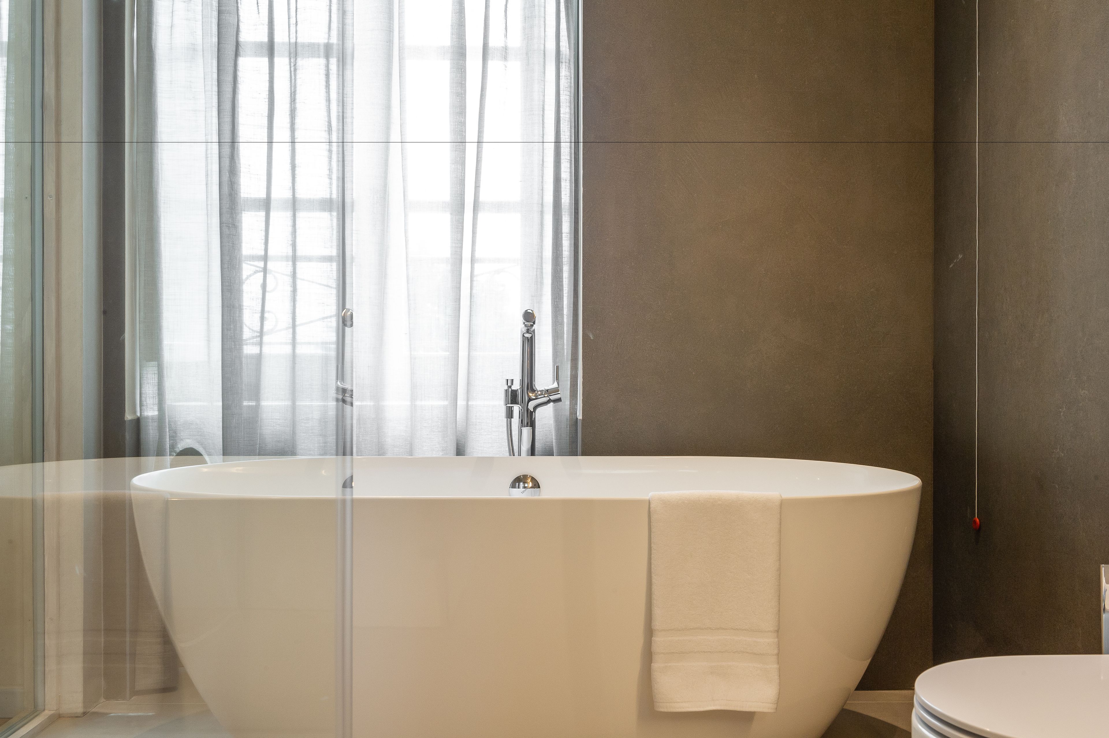 A modern bathroom with a freestanding bathtub, a toilet, and a large window with sheer curtains.