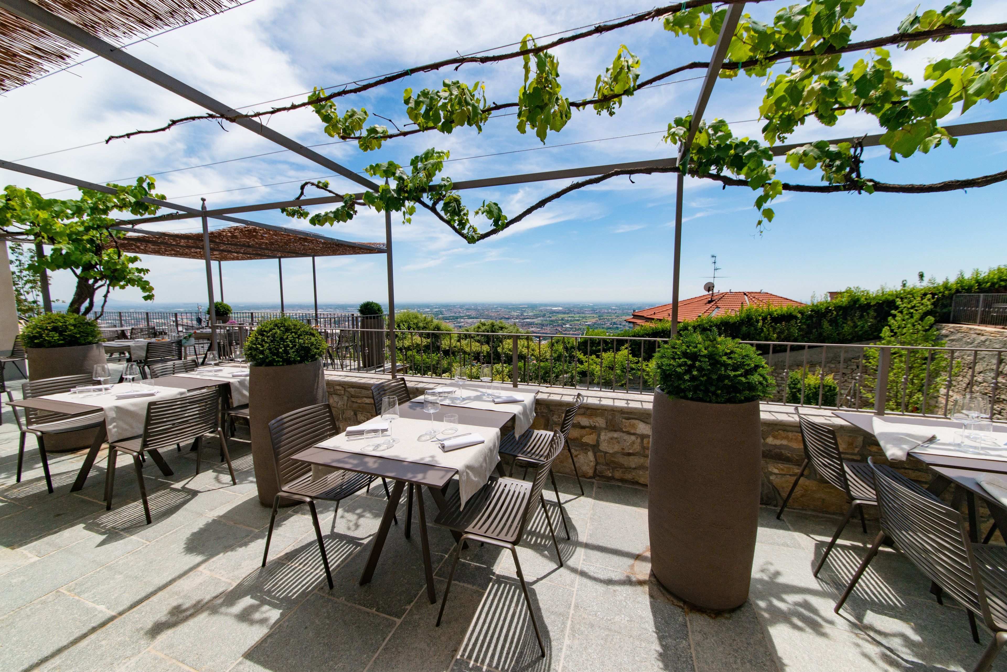 Eine Terrasse im Freien mit Tischen und Stühlen, umgeben von Steinmauern und Blumentöpfen, mit Blick auf eine schöne Aussicht.