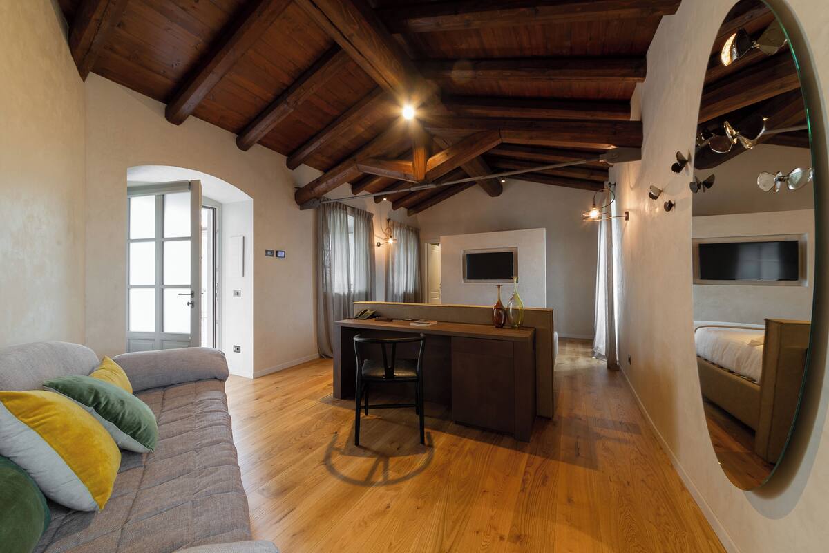 A spacious living area with a wooden ceiling, a sofa with cushions, a dining table with chairs, and a bed in the background.