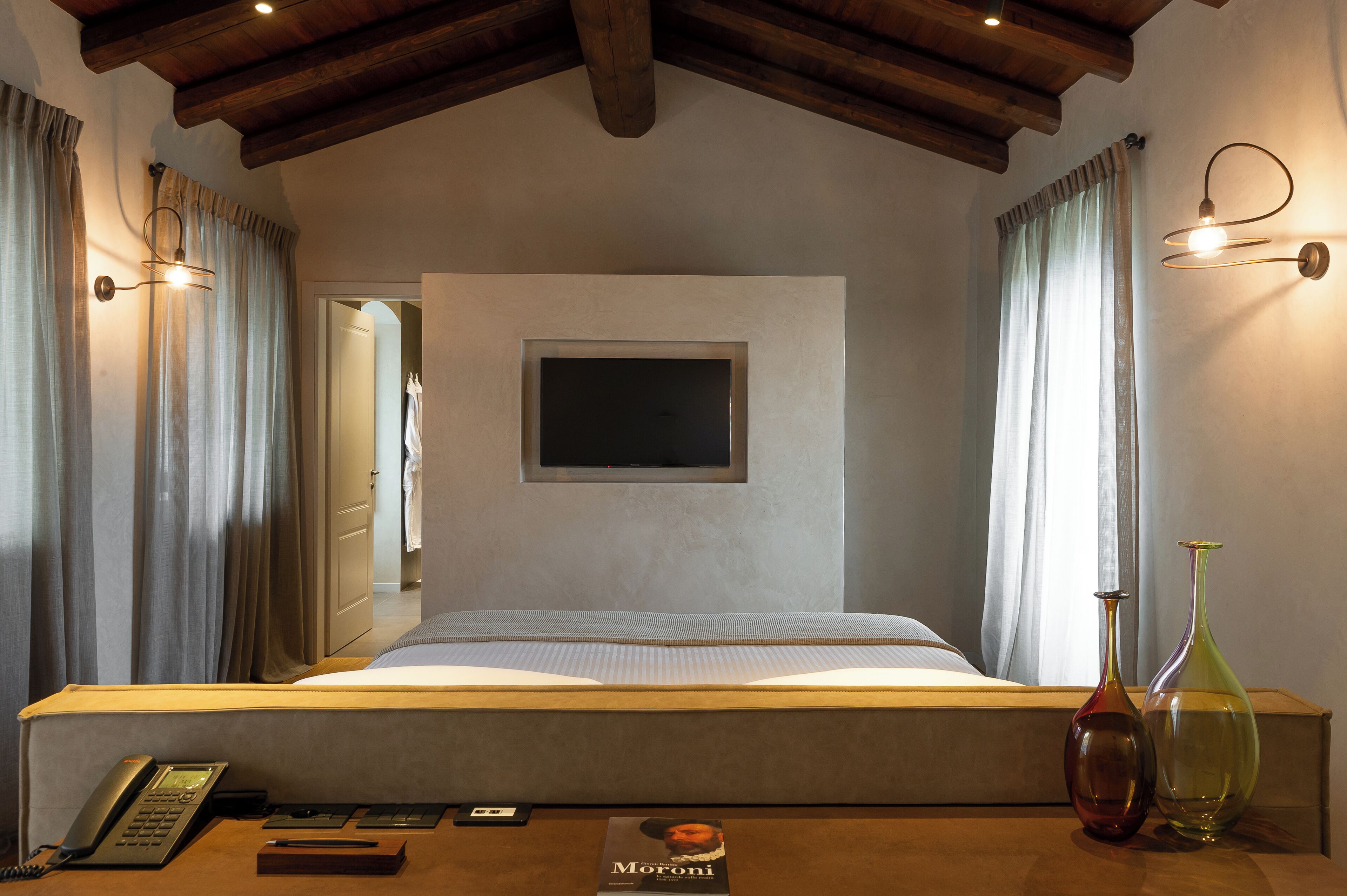 A bedroom with a bed, bedside table, lamp, and a flat-screen TV mounted on the wall.