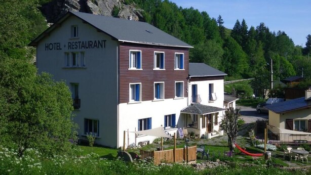 Ein Hotel mit Restaurant, das ein zweistöckiges Gebäude mit weißer Fassade und braunen Verzierungen inmitten üppiger Vegetation und eines felsigen Hügels zeigt.